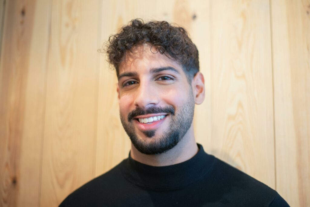Portrait of a smiling man indoors with curly hair and beard against a wooden backdrop.
